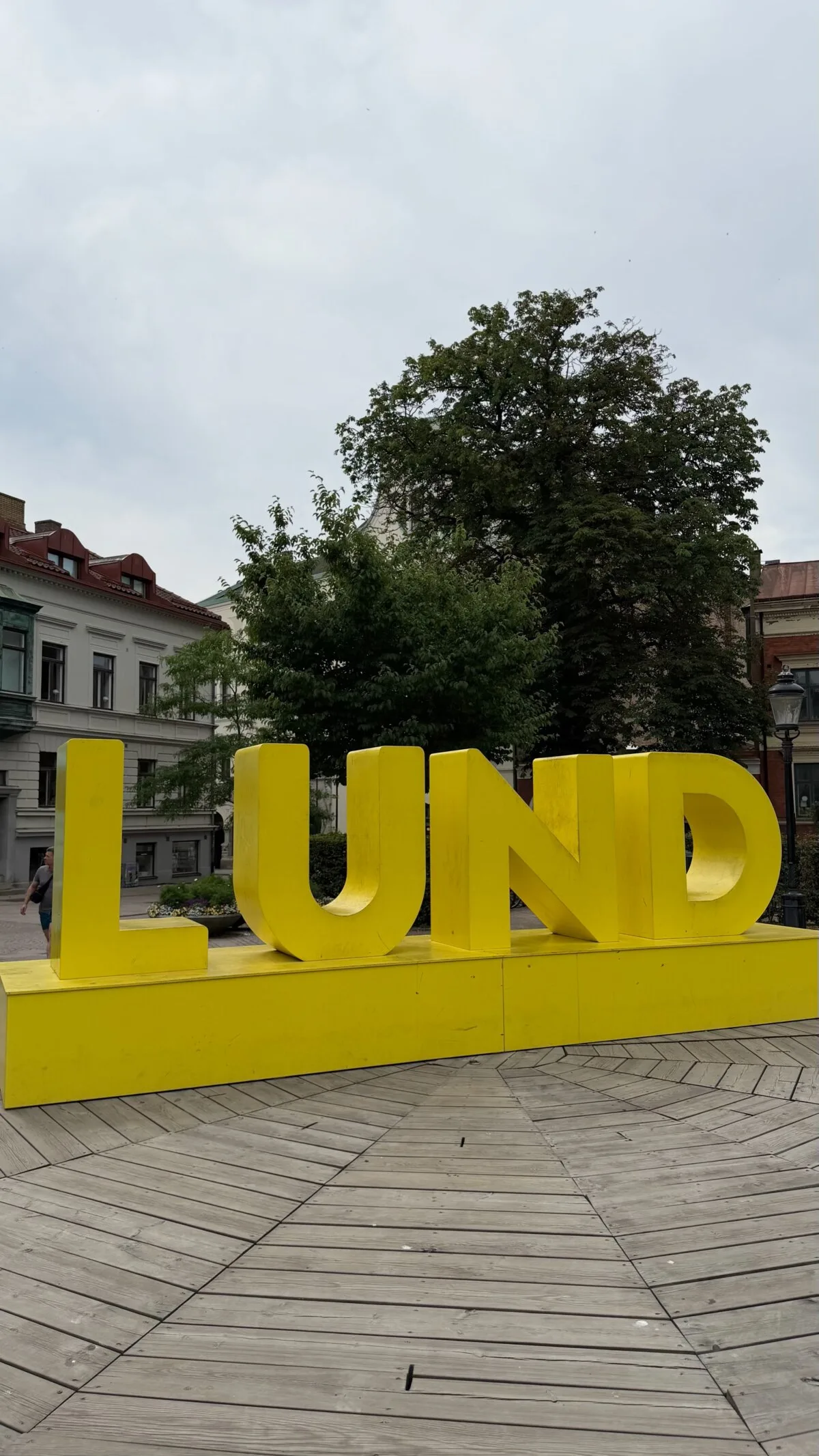 Giant yellow LUND letters on a square in Lund, Sweden, with brick buildings and trees behind
